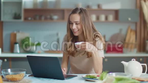 女人在摄像头上和朋友在厨房的笔记本电脑上聊天女士喝咖啡视频的预览图