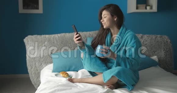 妇女在床上喝咖啡和使用智能手机视频的预览图