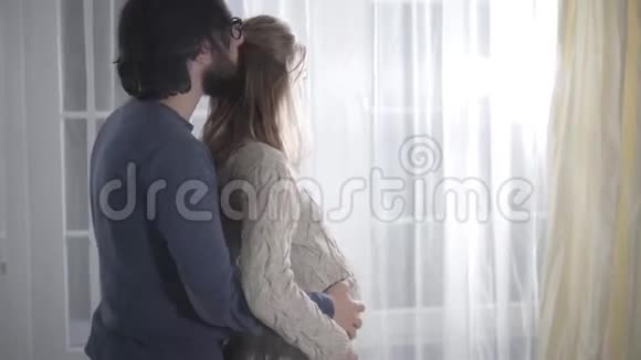 侧观留着胡子的白种人黑发男人亲吻妻子的头拥抱她怀孕的腹部幸福家庭视频的预览图