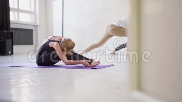 穿着黑色运动服的迷人柔韧女性在瑜伽室锻炼在垫子上伸展背部和腿部视频的预览图