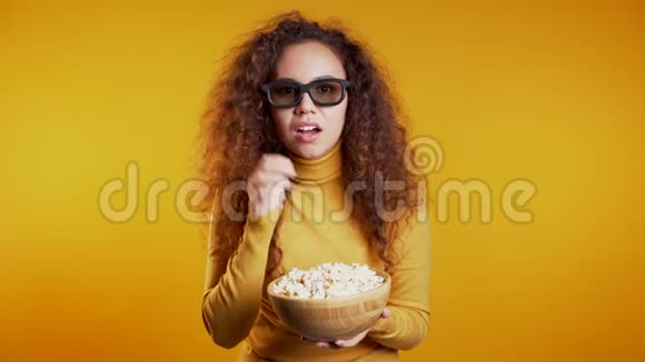 年轻女子在3d眼镜看电影和吃爆米花的黄色工作室背景视频的预览图