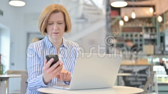 带手提电脑的创意老女人在咖啡厅用智能电话交谈视频的预览图