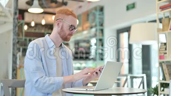 带手提电脑的红发男子在咖啡厅智能电话交谈视频的预览图