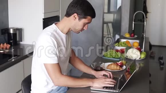 一个黑发年轻人早上坐在厨房的柜台上用笔记本电脑工作查看电子邮件社交媒体的人视频的预览图