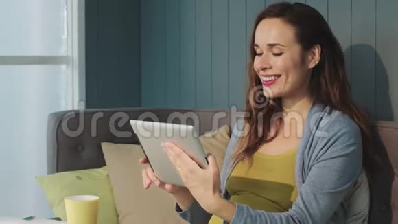微笑的孕妇坐在室内沙发上用平板电脑的肖像视频的预览图