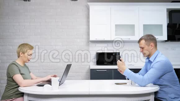 现代的家庭主妇和商人坐在厨房里互相忽视视频的预览图