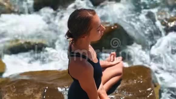 镜头右上方是穿着泳装盘腿坐在石头上的苗条年轻女子视频的预览图