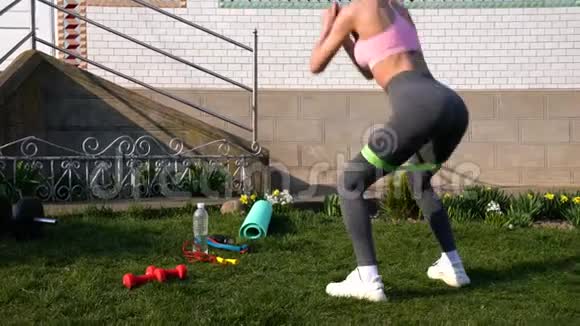 穿着运动服的年轻苗条女子在家里用橡皮筋在草地上做健身运动户外训练视频的预览图