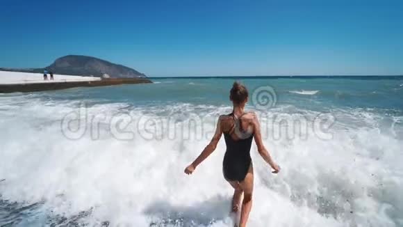 镜头跟随着穿着泳装的幸福苗条女人欢快地跑进挥舞的大海视频的预览图