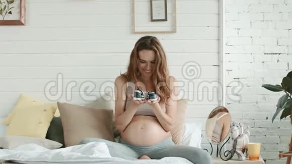 准妈妈在床上欣赏婴儿鞋微笑的孕妇白色房间视频的预览图