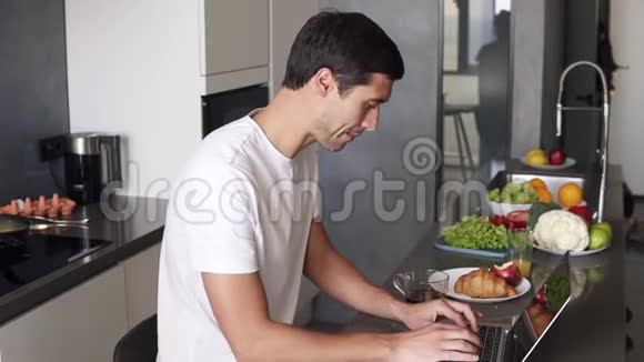 英俊的年轻人早上坐在厨房柜台的家里用笔记本电脑工作男人查看电子邮件社交媒体视频的预览图