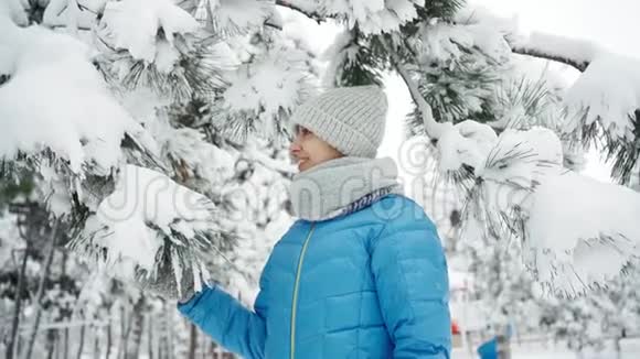 可爱的微笑的年轻女子穿着温暖的羊毛帽和长长的围巾在毛茸茸的一天在下雪的冬天公园里摇动着一根雪树枝视频的预览图