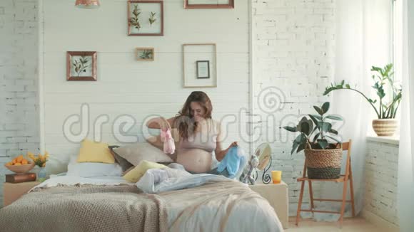 快乐的肚子妈妈看着婴儿衣服开心怀孕坐卧室视频的预览图