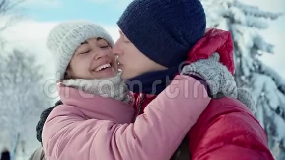 喜笑颜开的年轻夫妇在阳光明媚的冬天在雪地冬季公园散步时亲吻拥抱的特写镜头视频的预览图