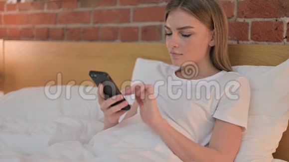 在床上使用智能手机庆祝成功的年轻女性视频的预览图