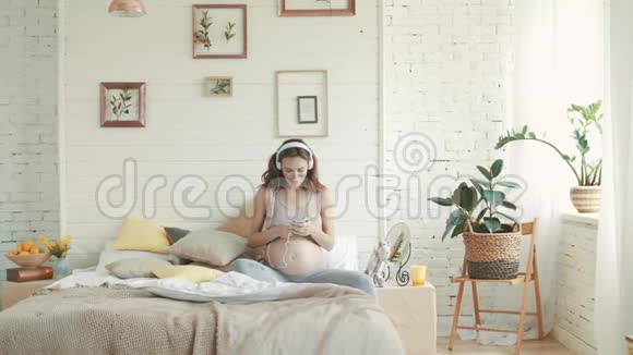 孕妇在床上听音乐准妈妈带着耳机坐着视频的预览图