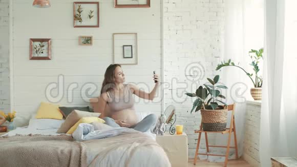 开朗的准妈妈拍自拍照片坐在床上的孕妇视频的预览图