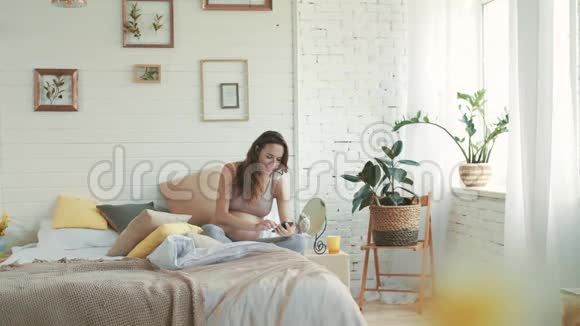 孕妇在床上拍自拍手机年轻的准妈妈坐在家里视频的预览图