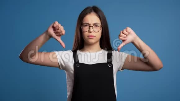 年轻女孩站在蓝工作室的背景下表达不满和拇指向下的手势视频的预览图