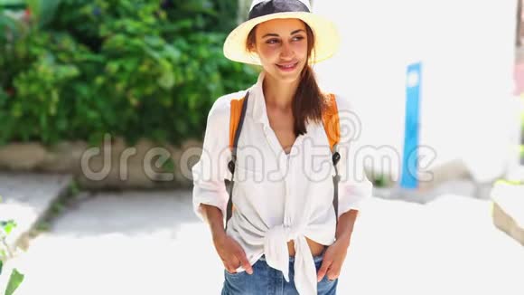 带着草帽白衬衫和橙色背包的迷人微笑的女游客在老城区的大街上散步视频的预览图