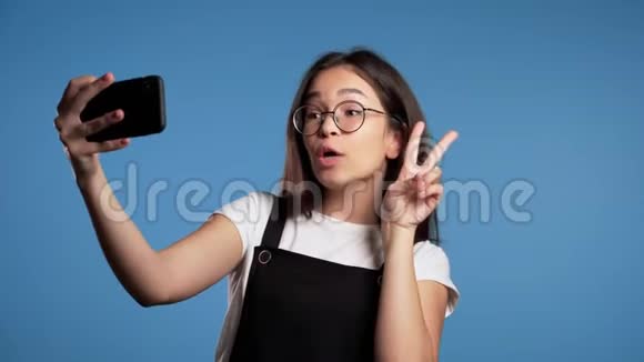 微笑快乐的亚洲女孩戴眼镜在智能手机上自拍蓝色背景视频的预览图
