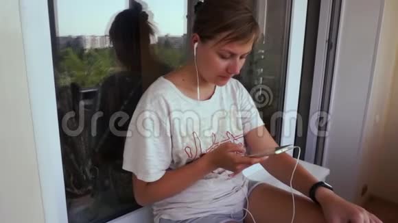 女孩听着智能手机的音乐戴着耳机在阳台上窗台上视频的预览图
