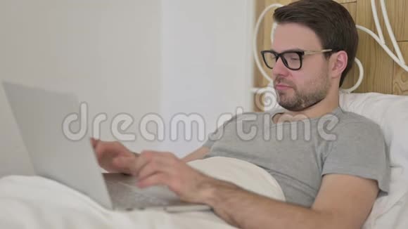 胡子青年男子在床上的笔记本电脑上得到惊喜视频的预览图