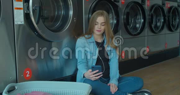 年轻的女人坐在地板上等待洗衣服在自助洗衣店里使用智能手机视频的预览图