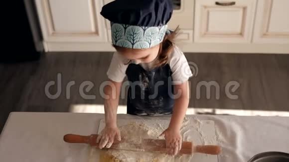 一个穿着厨师帽和围裙可爱的小女孩在家里卷着一个擀面杖面团视频的预览图