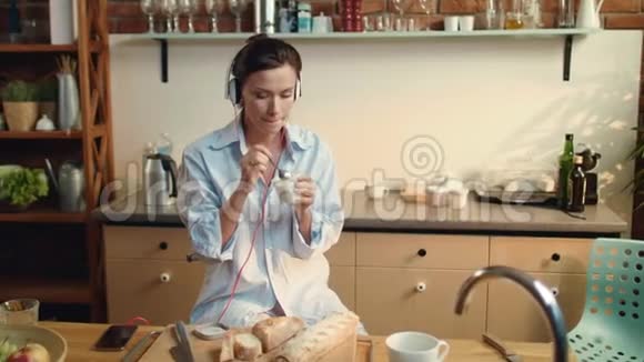 女人在厨房吃酸奶漂亮的女孩在耳机里听音乐视频的预览图