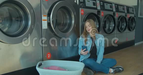 年轻女子在自助服务中坐在地板上听音乐等着洗衣服视频的预览图