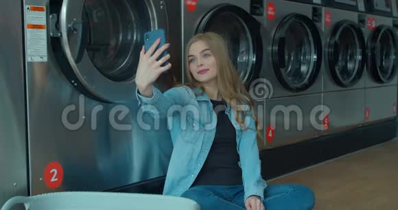 快乐的年轻女人在自助洗衣店的智能手机上自拍自助公共洗衣店视频的预览图