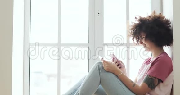 女人正在使用坐在窗口附近的智能手机制作在线支付银行卡视频的预览图