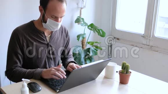 在家工作时戴着医用口罩的年轻人因为冠状病毒爆发在木桌上放了笔记本电脑和手机视频的预览图