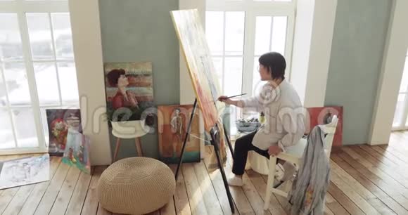 画家在画架上画画坐在靠近窗户的艺术工作室里视频的预览图