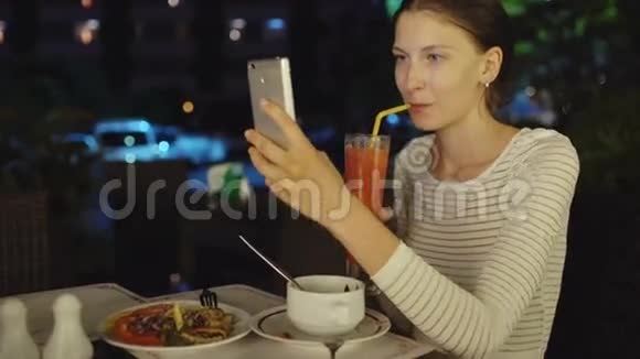 快乐的女人用智能手机和drinck鸡尾酒汁拍摄自拍晚上坐在餐厅里视频的预览图