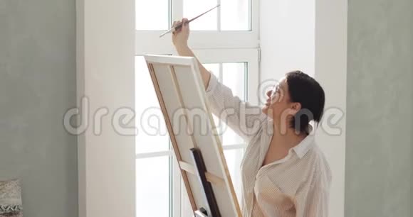 女画家正在画架上画画站在车间的窗户附近视频的预览图