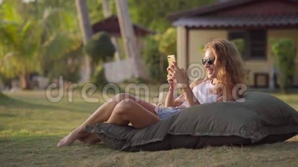 美丽的女孩躺在草地上的包椅上用智能手机工作视频的预览图