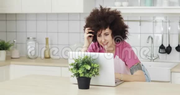 年轻的微笑的女人卷发在厨房的笔记本电脑上和朋友聊天视频的预览图