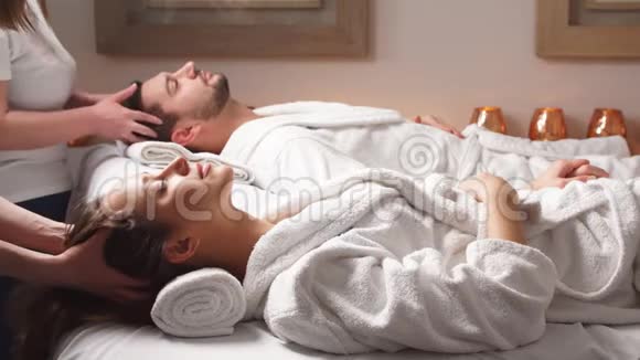 美丽的夫妇躺在按摩床上享受豪华水疗中心的头部按摩视频的预览图