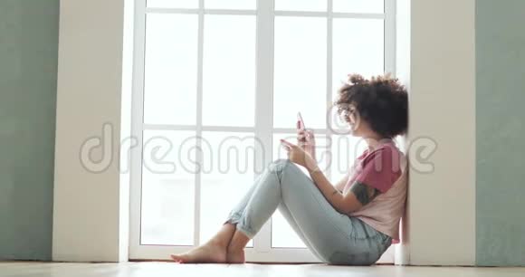 女孩正在使用坐在窗口附近的智能手机制作在线支付银行卡视频的预览图