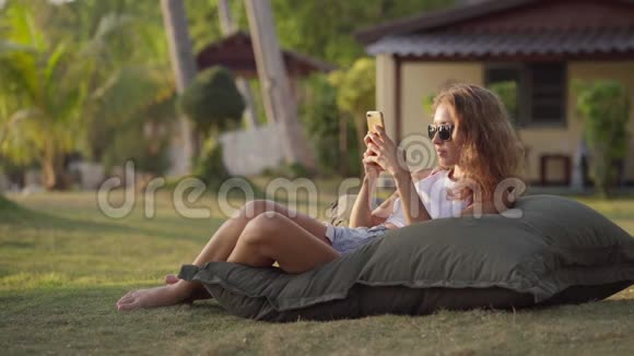 美丽的女孩躺在草地上的包椅上用智能手机工作视频的预览图