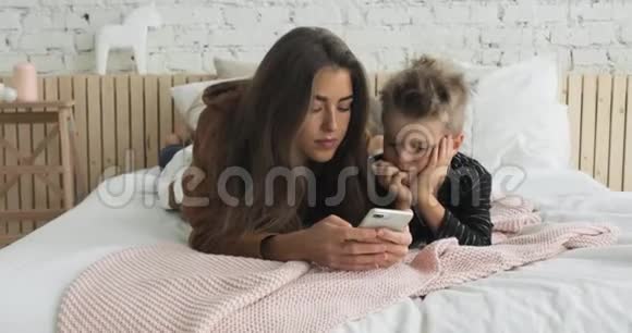 妈妈和儿子在托儿所的床上浏览智能手机孩子很无聊视频的预览图
