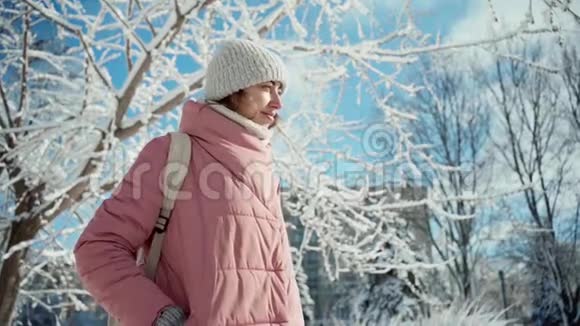 慢镜头美丽的微笑着的年轻女子穿着温暖的粉色派克大衣在阳光灿烂的白雪冬日公园里散步视频的预览图