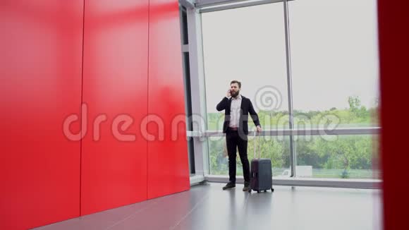 布吕内特蓄着胡须的男子正拿着一个带轮子的手提箱站在全景窗户旁用电话交谈然后离开视频的预览图