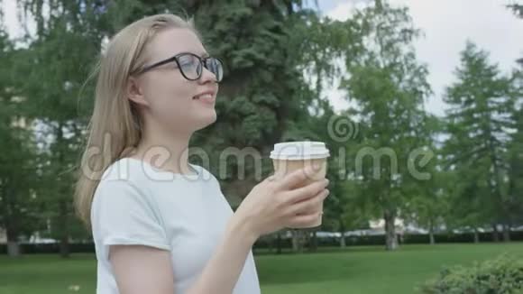 一个体贴的金发女郎戴着眼镜从外卖的杯子里喝着酒两只手抱着它梦幻地看着远离视频的预览图