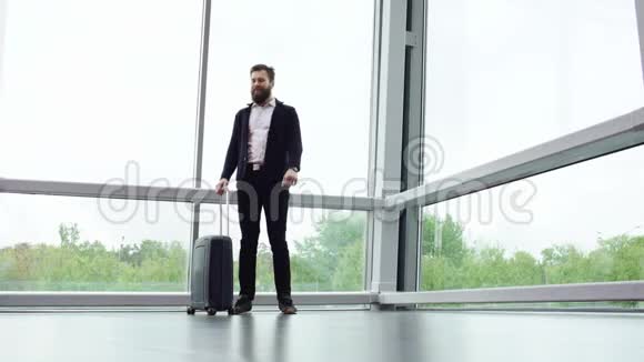 身着休闲装的长胡子黑发男子站在全景窗旁手提箱在轮子上看着手表视频的预览图