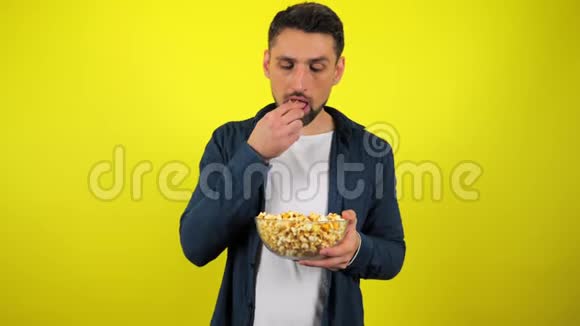 一个穿着蓝色衬衫和白色T恤的年轻人吃透明玻璃盘子里的爆米花并喜欢它视频的预览图