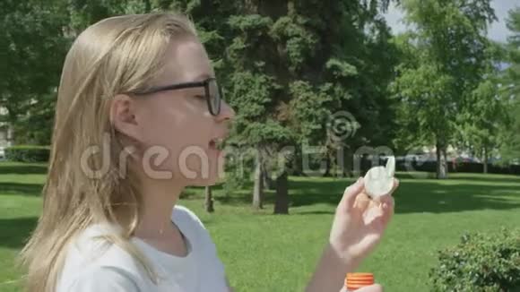 可爱的年轻女性金发天真地试图在绿色草坪前吹大肥皂泡侧视视频的预览图