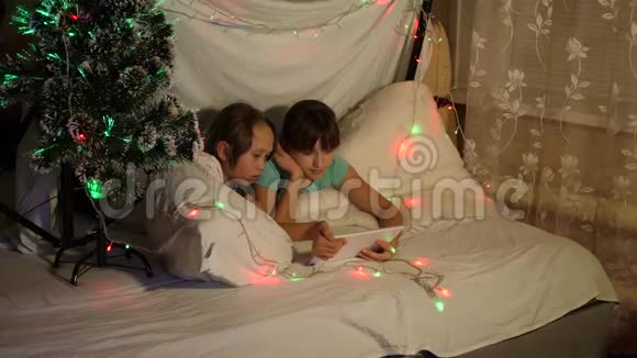 家庭圣诞节的概念孩子们在圣诞节晚上玩平板电脑在孩子们的房间里沙发上的花环s视频的预览图